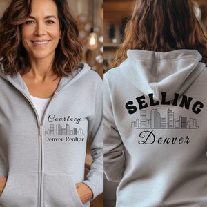Puede incluir: Una sudadera con capucha gris con cremallera y un gr&aacute;fico negro de un horizonte de ciudad y el texto "Selling Denver" en la parte posterior. La parte delantera de la sudadera tiene un gr&aacute;fico negro de un horizonte de ciudad y el texto "Courtney Denver Realtor".