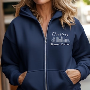 Puede incluir: Una sudadera con capucha azul marino con cremallera, con un contorno blanco de un horizonte de ciudad y el texto "Courtney Denver Realtor" en la parte delantera.