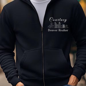 Puede incluir: Una sudadera con capucha negra con cremallera y un dise&ntilde;o blanco con un horizonte y el texto "Courtney Denver Realtor".