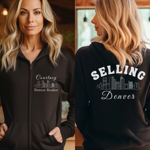 Puede incluir: Sudadera negra con cremallera y capucha, con un gr&aacute;fico blanco de un horizonte de ciudad y el texto "Selling Denver" en la parte posterior. La parte delantera de la sudadera tiene un gr&aacute;fico blanco de un horizonte de ciudad y el texto "Courtney Denver Realtor".