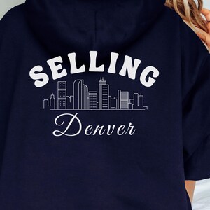 Puede incluir: Sudadera con capucha azul marino con un gr&aacute;fico blanco de un horizonte de ciudad y el texto "SELLING Denver".