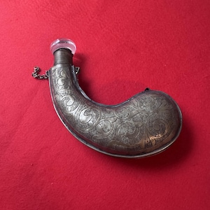 May include: A curved, metallic powder flask featuring intricate, embossed scrollwork patterns across its surface. The flask has a narrow, threaded neck topped with a clear, rounded glass stopper and includes a small metal chain attached to the side.