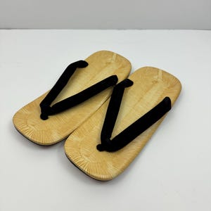 May include: A pair of traditional Japanese sandals, known as geta, with wooden soles and black fabric straps. The sandals have a light brown color and a textured pattern on the soles.
