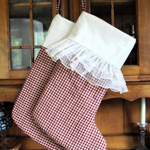 Puede incluir: Dos calcetines navideños con un patrón de cuadros vichy en rojo y blanco. Cada calcetín tiene un puño blanco sólido y un detalle de volantes en tela blanca transparente. Los calcetines están suspendidos por lazos de tela vichy a juego.