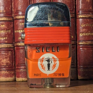 May include: A vintage orange and silver lighter with the word "SICLI" printed on the front. The lighter also has the words "PARIS" and "737 99 91" printed on the front.