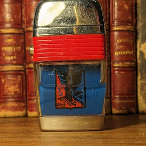 May include: A silver and red lighter with a blue and red eagle design and the number 453. The lighter is sitting on a wooden surface.