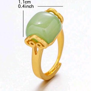 May include: A gold-colored ring featuring a light green, oval-shaped stone. The stone measures 0.4 inches in length. The ring has an ornate design around the stone and a simple band.