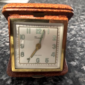 May include: A vintage travel alarm clock with a brown leather case and a square face. The clock face is white with black numerals and gold hands. The clock is closed, with the top cover in place.