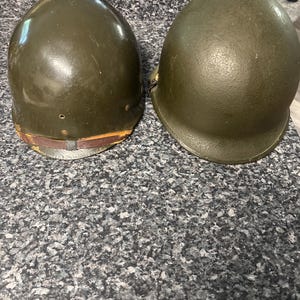 May include: Two olive drab military helmets. The helmet on the left has a smooth, glossy finish with a brown leather chin strap attached. The helmet on the right features a textured, matte finish and a rounded dome shape with a wide brim.