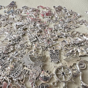 May include: A large collection of silver charm pendants featuring various designs, including owls, anchors, butterflies, and a wheel. The charms are perfect for jewelry making or crafting.