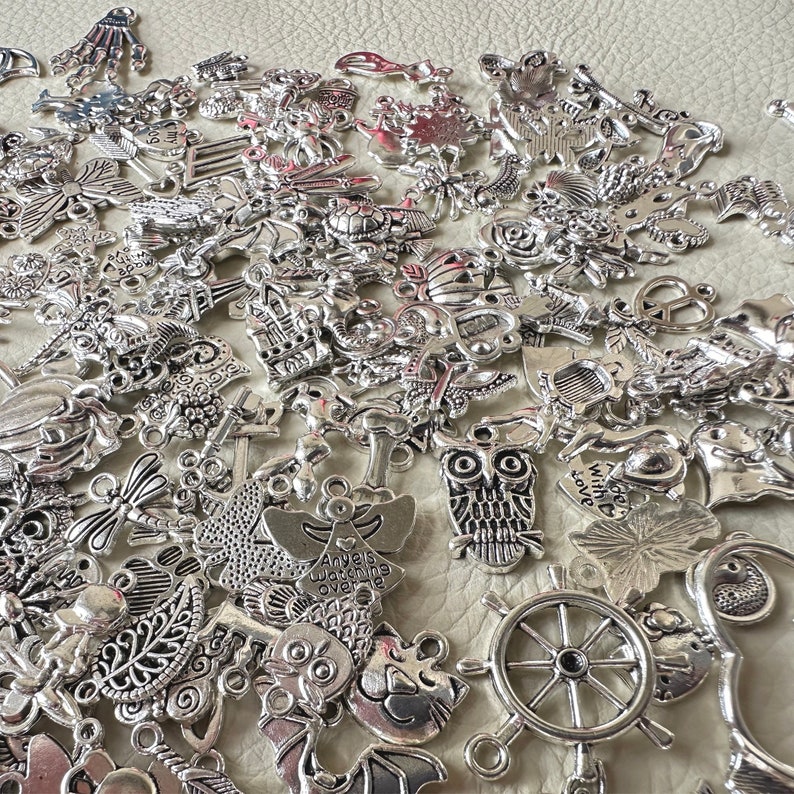 May include: A large collection of silver charm pendants featuring various designs, including owls, anchors, butterflies, and a wheel. The charms are perfect for jewelry making or crafting.