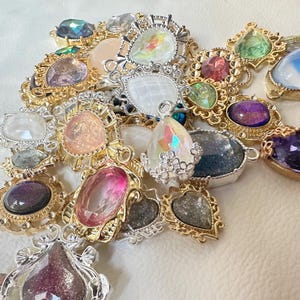 May include: A selection of decorative gemstone pendants in gold and silver settings. The pendants showcase a variety of shapes and colours, including pink, purple, green, and iridescent stones. Ideal for jewellery making or craft projects.