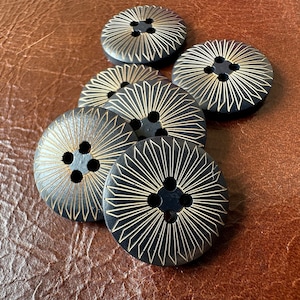 May include: Six black buttons with a gold sunburst pattern. Each button has four holes for sewing. The buttons are circular and made of a dark material. The buttons are on a brown leather-like surface.