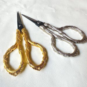 May include: Two pairs of decorative scissors. One pair has gold-colored handles with a detailed, ornate design. The other pair has silver-colored handles with a similar design. Both pairs have black blades.