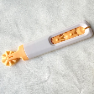 May include: A white and orange pastry cutter with a decorative wheel and several interchangeable shapes. The tool is designed for cutting dough and creating decorative edges on pastries and baked goods.