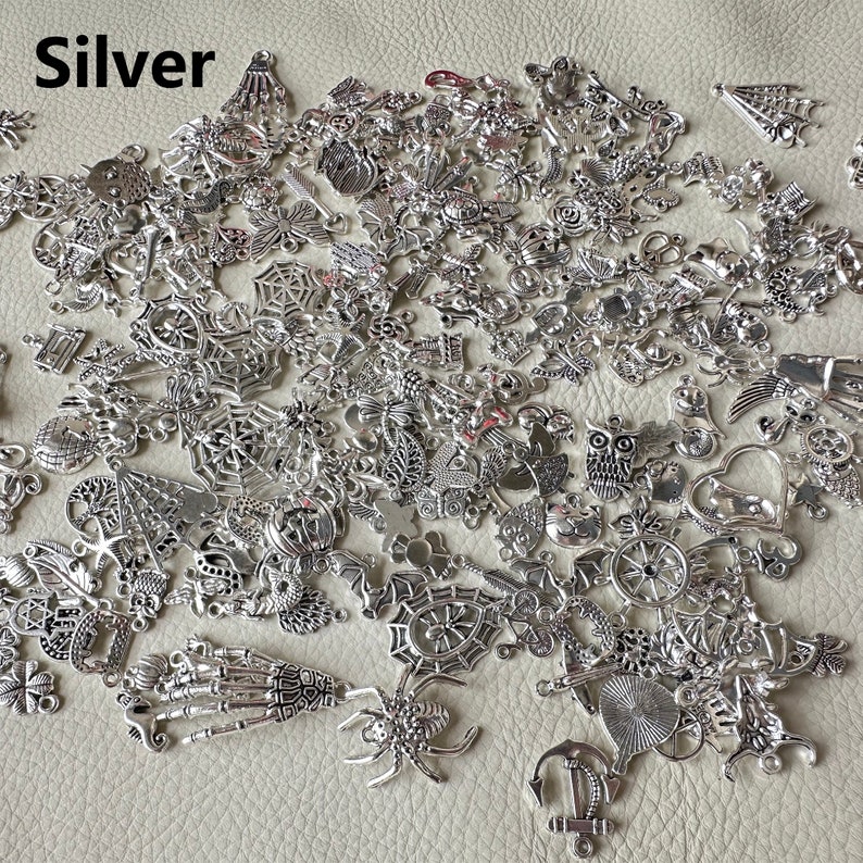 May include: A large assortment of silver charm pendants in various shapes and designs, including spiders, anchors, hearts, owls, and more. The charms are scattered on a light brown surface.