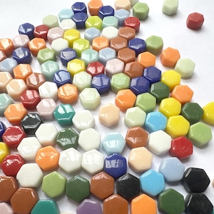Op de afbeelding: Een verzameling kleine, gladde, zeshoekige tegels in een grote verscheidenheid aan kleuren, waaronder rood, oranje, geel, groen, blauw, wit, zwart en beige. De tegels zijn verspreid over een effen wit oppervlak en hebben elk een glanzende afwerking.