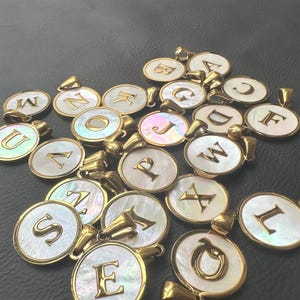 Op de afbeelding: Een verzameling goudkleurige initiaalhangers met iriserende parelmoer achtergronden. Elke hanger heeft een andere letter van het alfabet. De hangers zijn rond met een gouden rand en een lus voor een ketting.