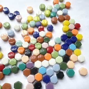 Pu&ograve; includere: Una collezione di piccole piastrelle esagonali lisce in un'ampia variet&agrave; di colori, tra cui blu, verde, arancione, giallo, bianco, nero, marrone e rosa. Le piastrelle sono sparse su una superficie bianca uniforme e presentano finiture lucide.