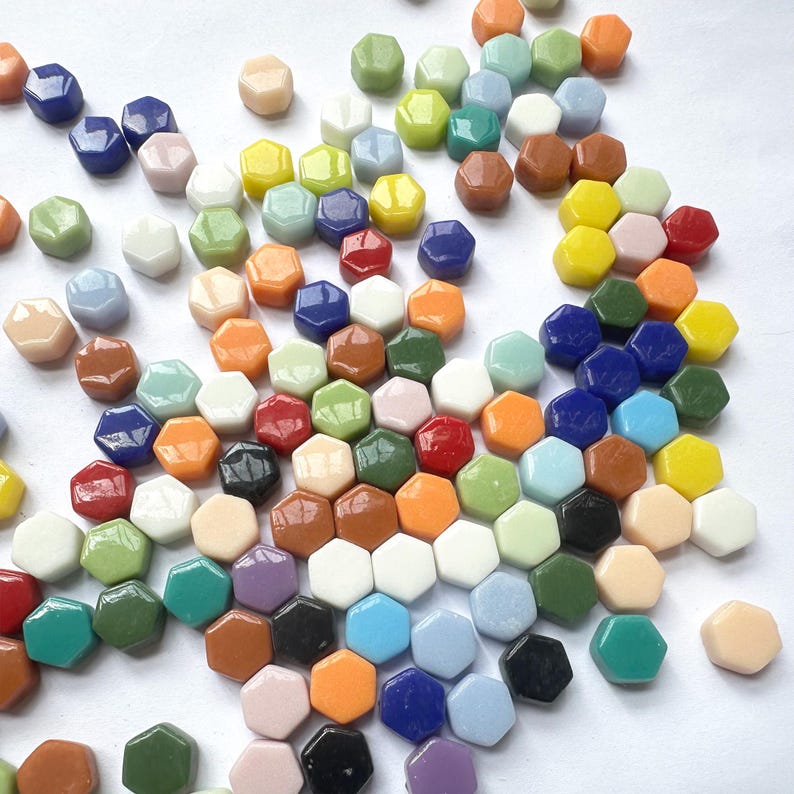 Pu&ograve; includere: Una collezione di piccole piastrelle esagonali lisce in un'ampia variet&agrave; di colori, tra cui blu, verde, arancione, giallo, bianco, nero, marrone e rosa. Le piastrelle sono sparse su una superficie bianca uniforme e presentano finiture lucide.
