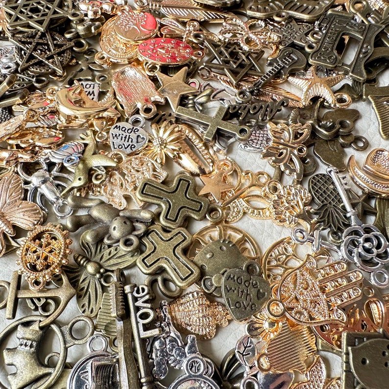 May include: A large collection of mixed metal charms in gold, silver, and bronze tones. The charms feature various designs, including crosses, stars, hearts, butterflies, and keys. Some charms have words on them, such as "Made with Love" and "Long".
