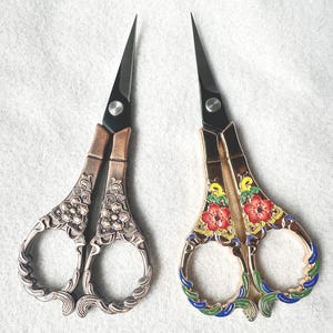 Könnte beinhalten: Zwei Paar dekorative Scheren. Ein Paar hat eine Bronzefinish mit floralen und Scrollwork-Details. Das andere Paar hat ein Gold-Finish mit roten, gelben und blauen floralen Designs. Beide haben schwarze Klingen und sind zum Basteln bestimmt.