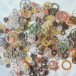 Op de afbeelding: Een verzameling metalen tandwielen van verschillende afmetingen in tinten zilver, goud, brons, koper en groen. De tandwielen zijn verspreid over een wit oppervlak, wat een steampunk- of industriële esthetiek creëert. Sommige tandwielen hebben ingewikkelde ontwerpen.