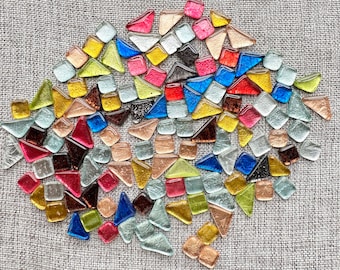 Può includere: Una collezione di piccole tessere di vetro colorate in varie forme, tra cui quadrati, triangoli e rettangoli. Le tessere sono disposte in uno schema casuale su una superficie testurizzata di colore marrone chiaro.