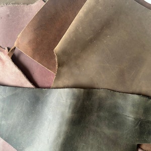 May include: A close-up of four pieces of leather in different shades of brown, gray, and purple. The leather is textured and has a worn look.