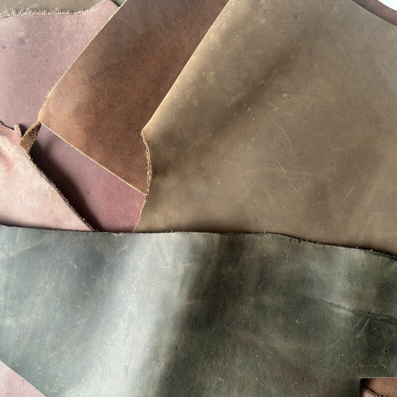 May include: A close-up of four pieces of leather in different shades of brown, gray, and purple. The leather is textured and has a worn look.