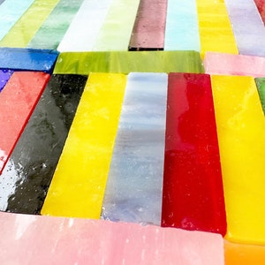 Op de afbeelding: Assortiment van kleurrijke glazen staven in verschillende tinten blauw, rood, geel, groen en wit. De glazen staven zijn in een rij gerangschikt, met de uiteinden van de staven die elkaar overlappen.