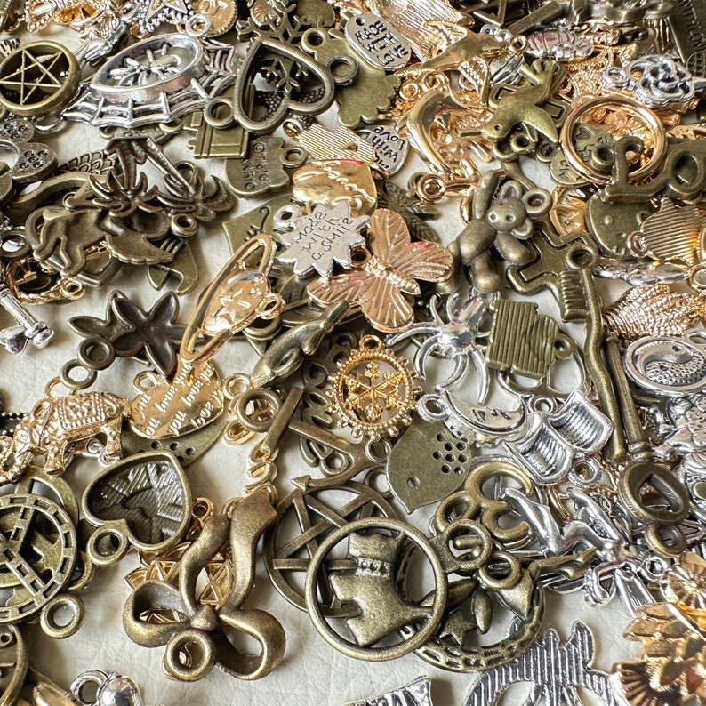 May include: A large collection of mixed metal charms in silver, gold, and bronze tones. The charms feature various designs, including hearts, stars, butterflies, and keys. Some charms have words or phrases on them, such as "Love you" and "Be Strong".