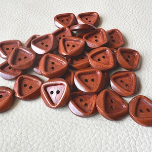 May include: A collection of triangular wooden buttons in a rich brown hue. Each button features two small holes for sewing. The buttons are scattered on a light-colored surface, showcasing their unique shape and texture.