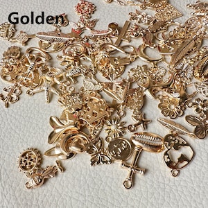 May include: A large assortment of gold-colored metal charms in various shapes, including hearts, stars, flowers, leaves, and more. The charms are scattered on a light beige surface.
