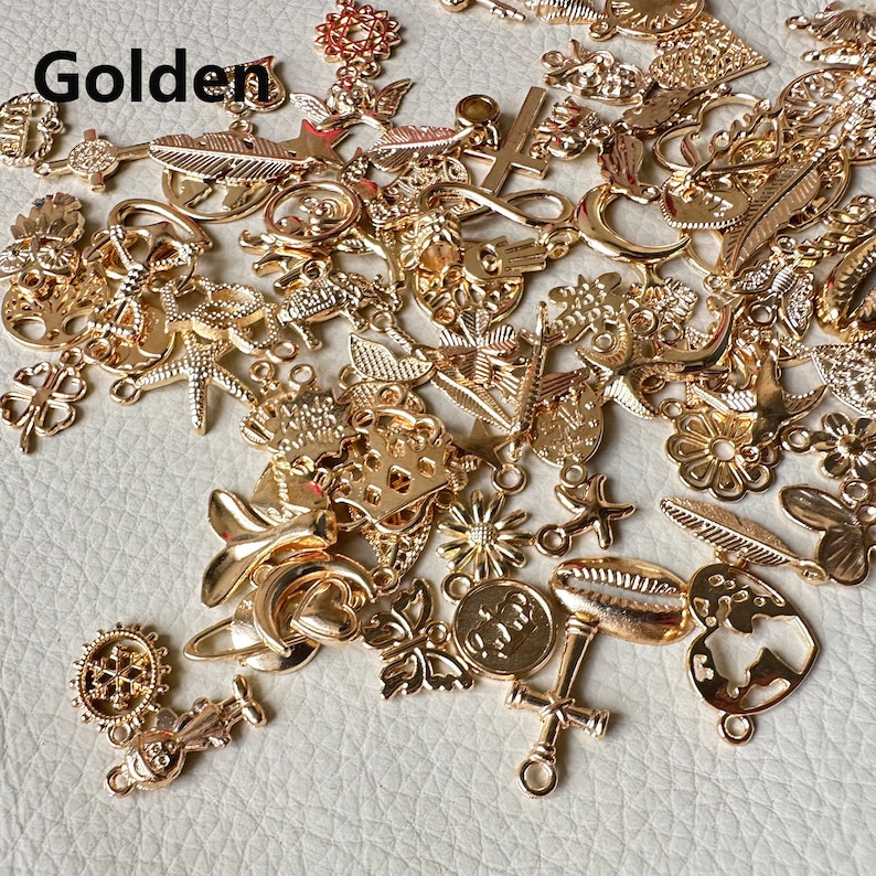 May include: A large assortment of gold-colored metal charms in various shapes, including hearts, stars, flowers, leaves, and more. The charms are scattered on a light beige surface.