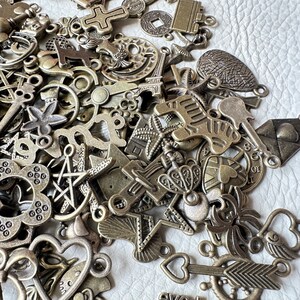 May include: A large collection of antique-looking metal charms in various shapes and sizes. The charms are made of a dark, aged metal and have a rustic, vintage look. Some of the charms include a cross, a star, a heart, a flower, and a bird.