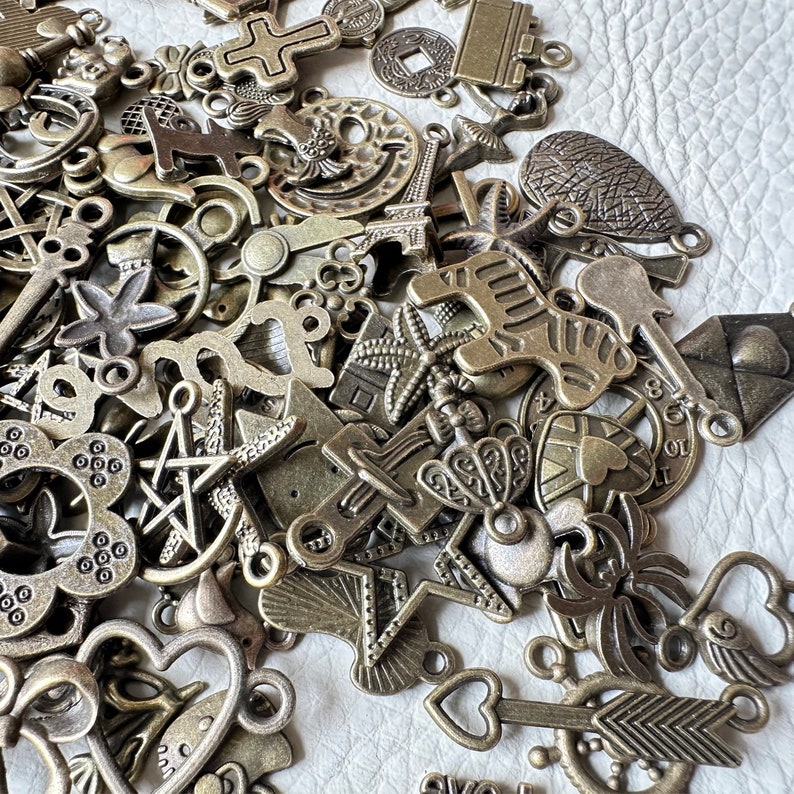 May include: A large collection of antique-looking metal charms in various shapes and sizes. The charms are made of a dark, aged metal and have a rustic, vintage look. Some of the charms include a cross, a star, a heart, a flower, and a bird.