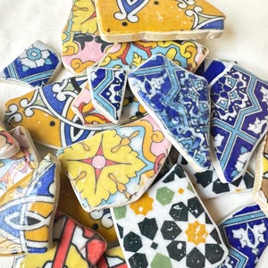 May include: A collection of broken ceramic tiles with colorful patterns. The tiles feature various designs in shades of blue, yellow, pink, and green. Some tiles have geometric shapes, while others have floral or abstract motifs. The pieces are arranged in a scattered composition.