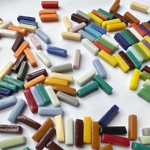 May include: A collection of small, rectangular glass mosaic tiles in a variety of colors including red, orange, yellow, green, blue, brown, white, and gray. The tiles are scattered across the surface, each featuring a smooth, glossy finish.