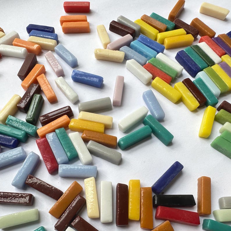 May include: A collection of small, rectangular glass mosaic tiles in a variety of colors including red, orange, yellow, green, blue, brown, white, and gray. The tiles are scattered across the surface, each featuring a smooth, glossy finish.