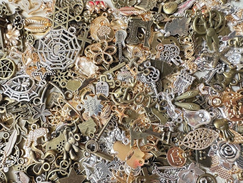 May include: A large collection of mixed metal charms in silver, gold, and bronze tones. The charms feature a variety of designs, including stars, crosses, hearts, flowers, and more.