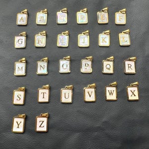 Puede incluir: Una colección de colgantes dorados, cada uno con una letra del alfabeto. Los colgantes tienen forma rectangular con una incrustación de nácar y un pequeño lazo en la parte superior para una cadena. Las letras están en una fuente serif clásica.