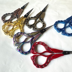 May include: A collection of decorative embroidery scissors in various colors, including gold, bronze, black, blue, red, and iridescent. The scissors have ornate handles and sharp, pointed blades, ideal for crafting and sewing projects.