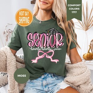 May include: A moss green short-sleeve t-shirt featuring a graphic print with the word SENIOR in pink bubble letters, a black graduation cap, the text twenty twenty-seven, and a pink ribbon bow graphic centered on the front of the garment.