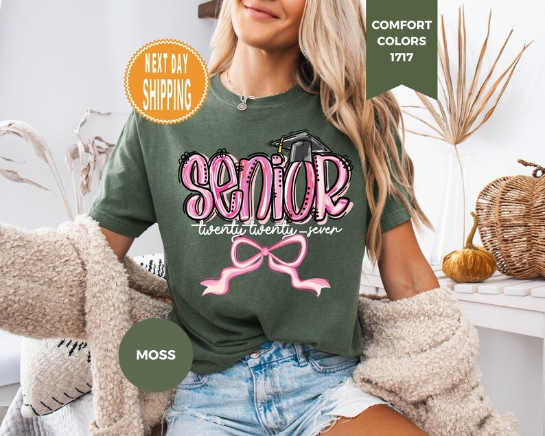 May include: A moss green short-sleeve t-shirt featuring a graphic print with the word SENIOR in pink bubble letters, a black graduation cap, the text twenty twenty-seven, and a pink ribbon bow graphic centered on the front of the garment.