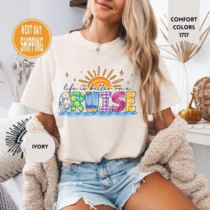 May include: A white t-shirt with a colorful graphic that says "Life is better on a cruise". The graphic features a sun, waves, and a colorful word "cruise".