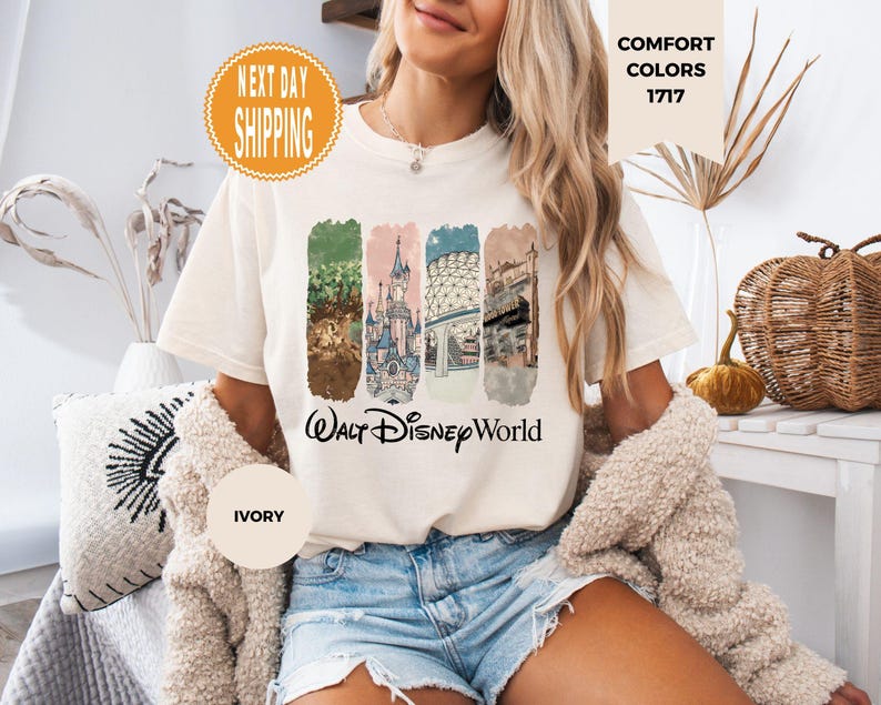 May include: Ivory-colored t-shirt featuring a colorful graphic of Walt Disney World landmarks. The design includes images of a castle, and other park features. The words "Walt Disney World" are printed below the graphic. The shirt has short sleeves and a crew neck.