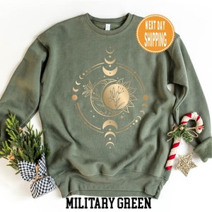 Op de afbeelding: Een militair groen sweatshirt met een gouden folie ontwerp van een zon, maan en sterren. De tekst "Military Green" is onderaan de afbeelding gedrukt.
