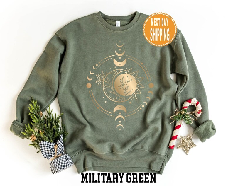 Op de afbeelding: Een militair groen sweatshirt met een gouden folie ontwerp van een zon, maan en sterren. De tekst "Military Green" is onderaan de afbeelding gedrukt.