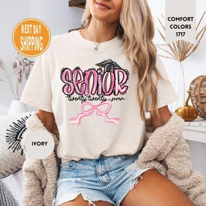 May include: An ivory short-sleeved t-shirt featuring a graphic with the word SENIOR in pink bubble letters, a black graduation cap, the text twenty twenty-seven, and a pink ribbon bow. The shirt includes a Comfort Colors 1717 label.